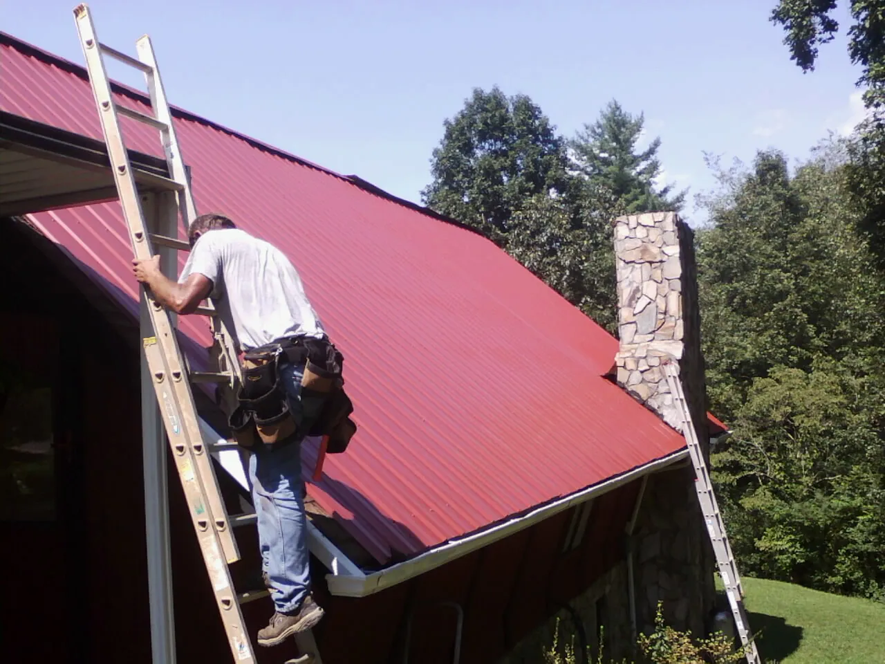 Roofer on a ladder beside a red metal roof and stone chimney in Pillager
